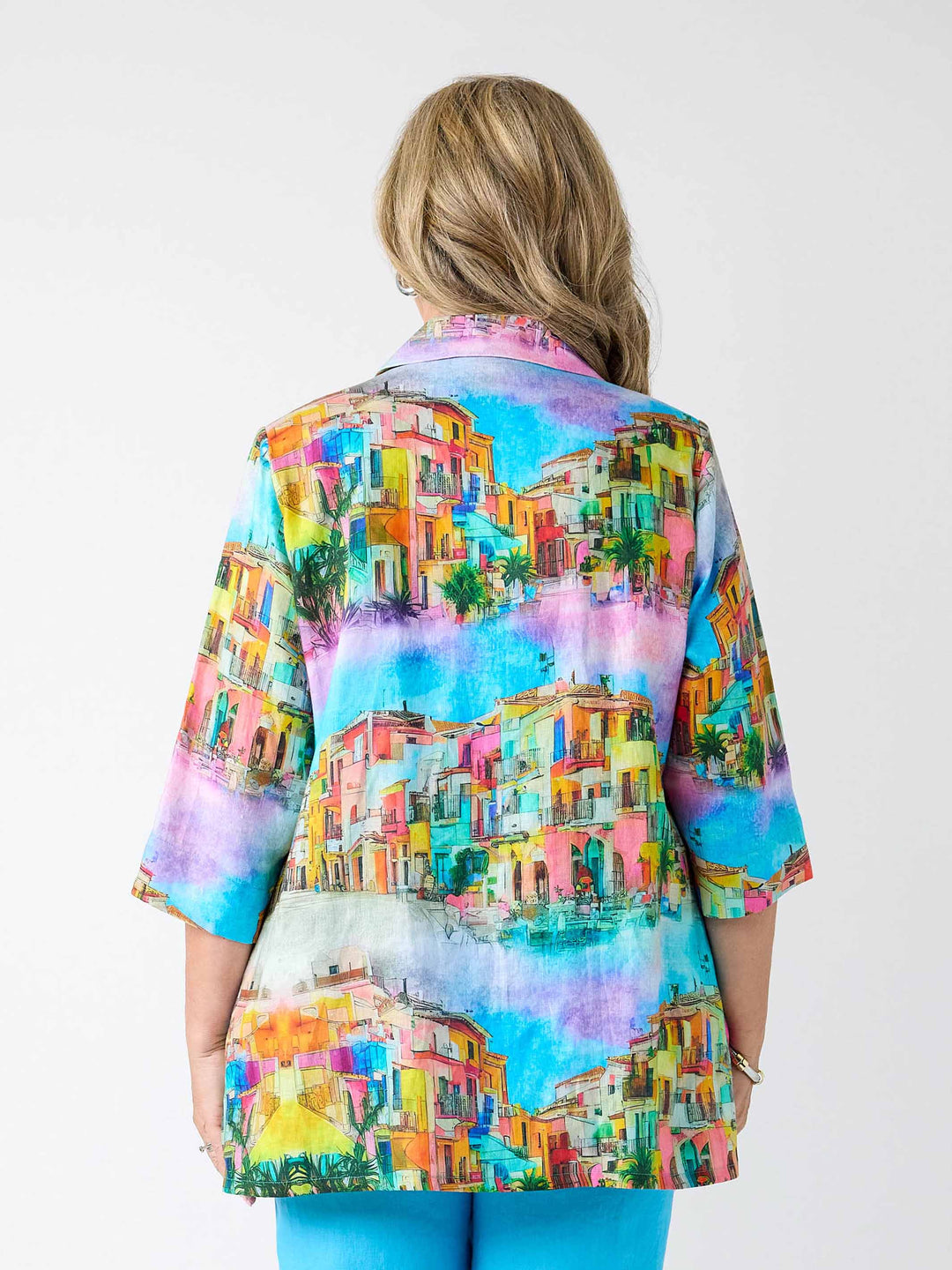A woman viewed from the back wearing a colourful short-sleeve button-up shirt with an all-over watercolor-style print of Mediterranean buildings in shades of blue, pink, yellow, orange, and green, paired with bright blue pants