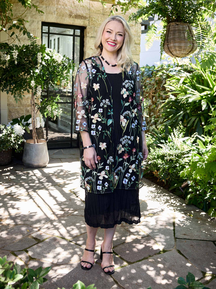 A woman wearing a sheer black embroidered evening jacket over a black crushed dress