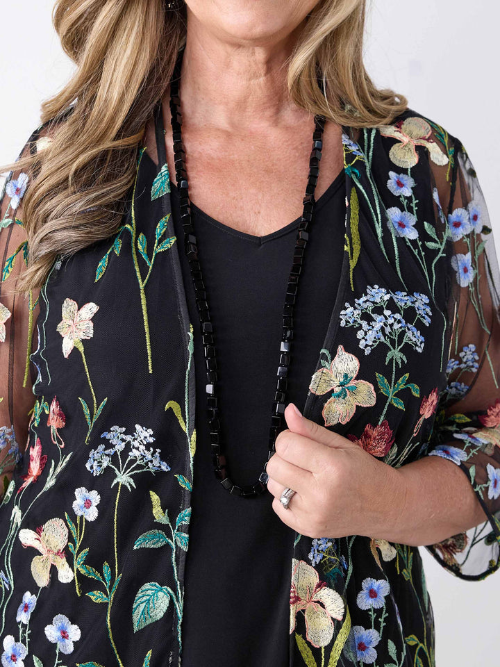 A close-up of a woman wearing a sheer black embroidered evening jacket over a black cami