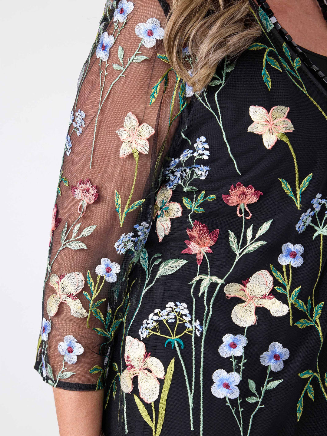 A close-up of a woman wearing a sheer black embroidered evening jacket over a black cami
