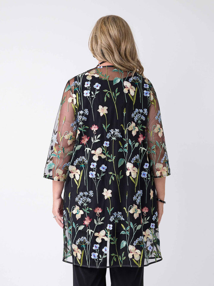 A back view of a woman wearing a sheer black embroidered evening jacket over a black cami