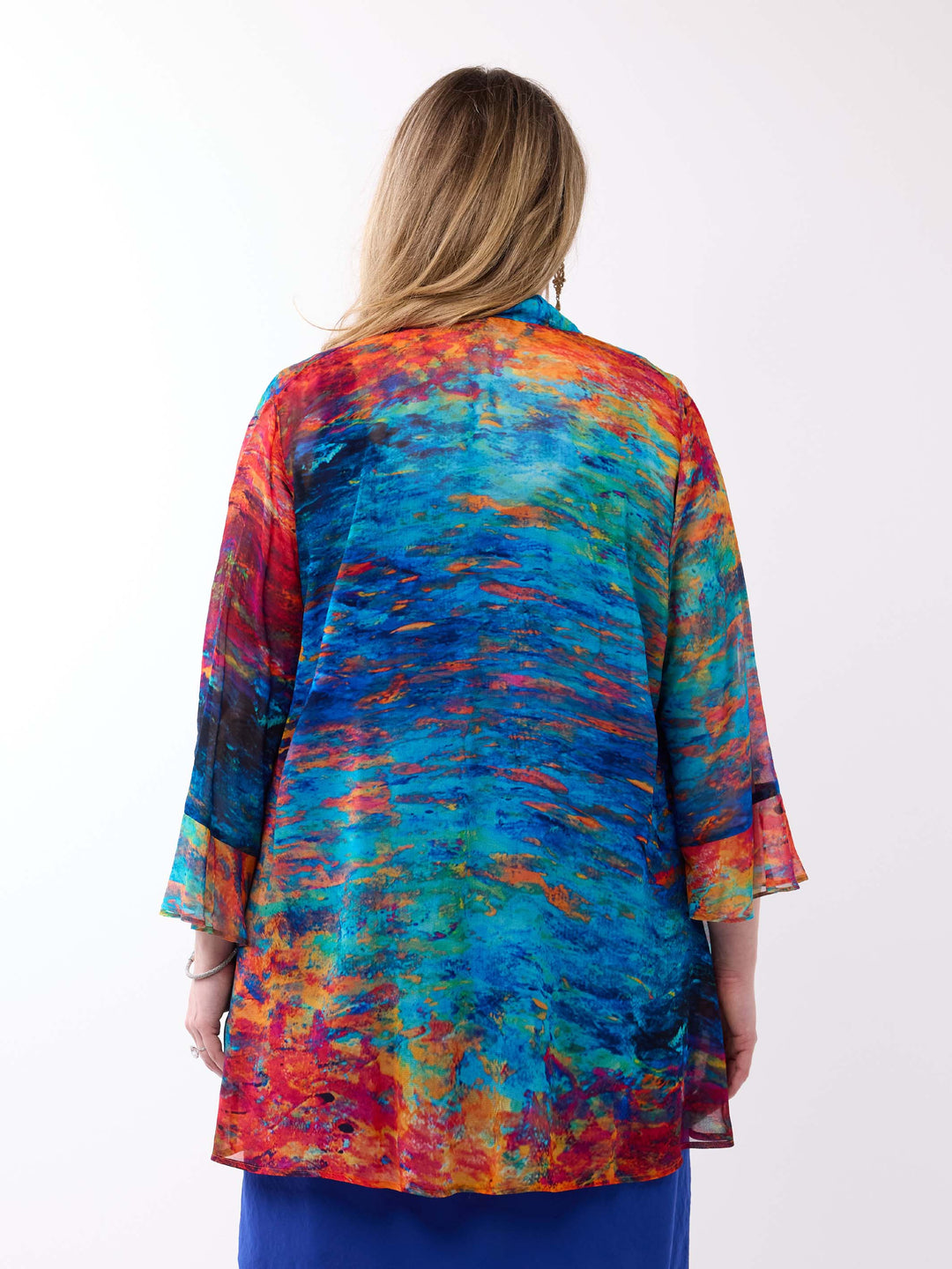 A woman viewed from behind wearing a vibrant, multicoloured abstract print top with blue, orange, red, and yellow tones, paired with blue dress