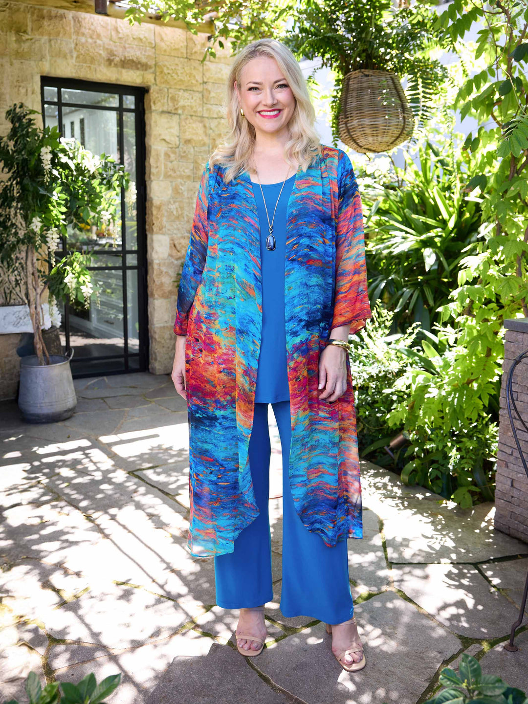 A woman wearing a vibrant, multicoloured abstract print long-line georgette evening jacket over a blue cami and palazzo pant. 
