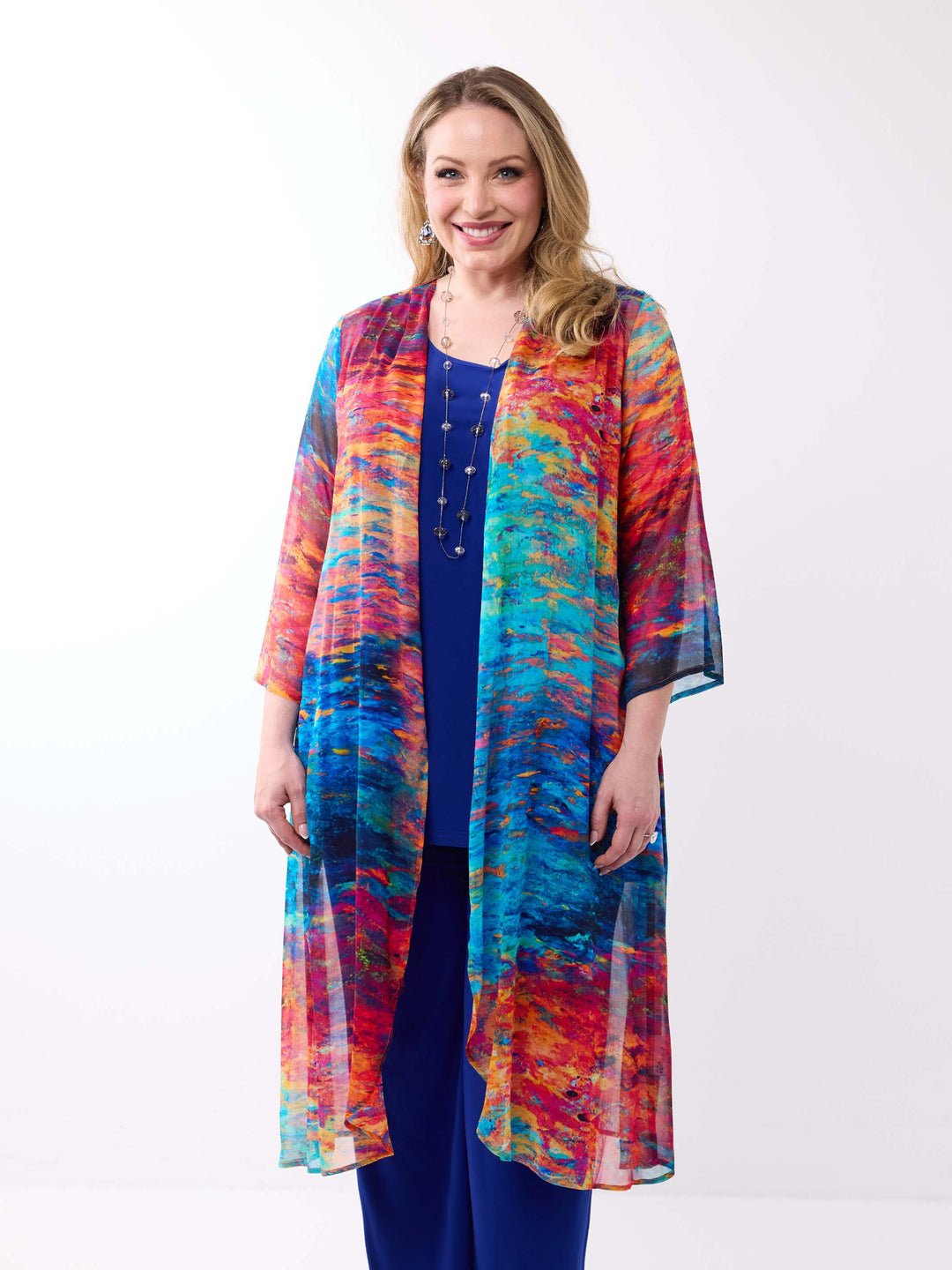 A woman wearing a lightweight, open-front long georgette evening jacket with a vibrant, multicolorued abstract print in shades of blue, red, orange, and yellow over a solid royal blue top and pants