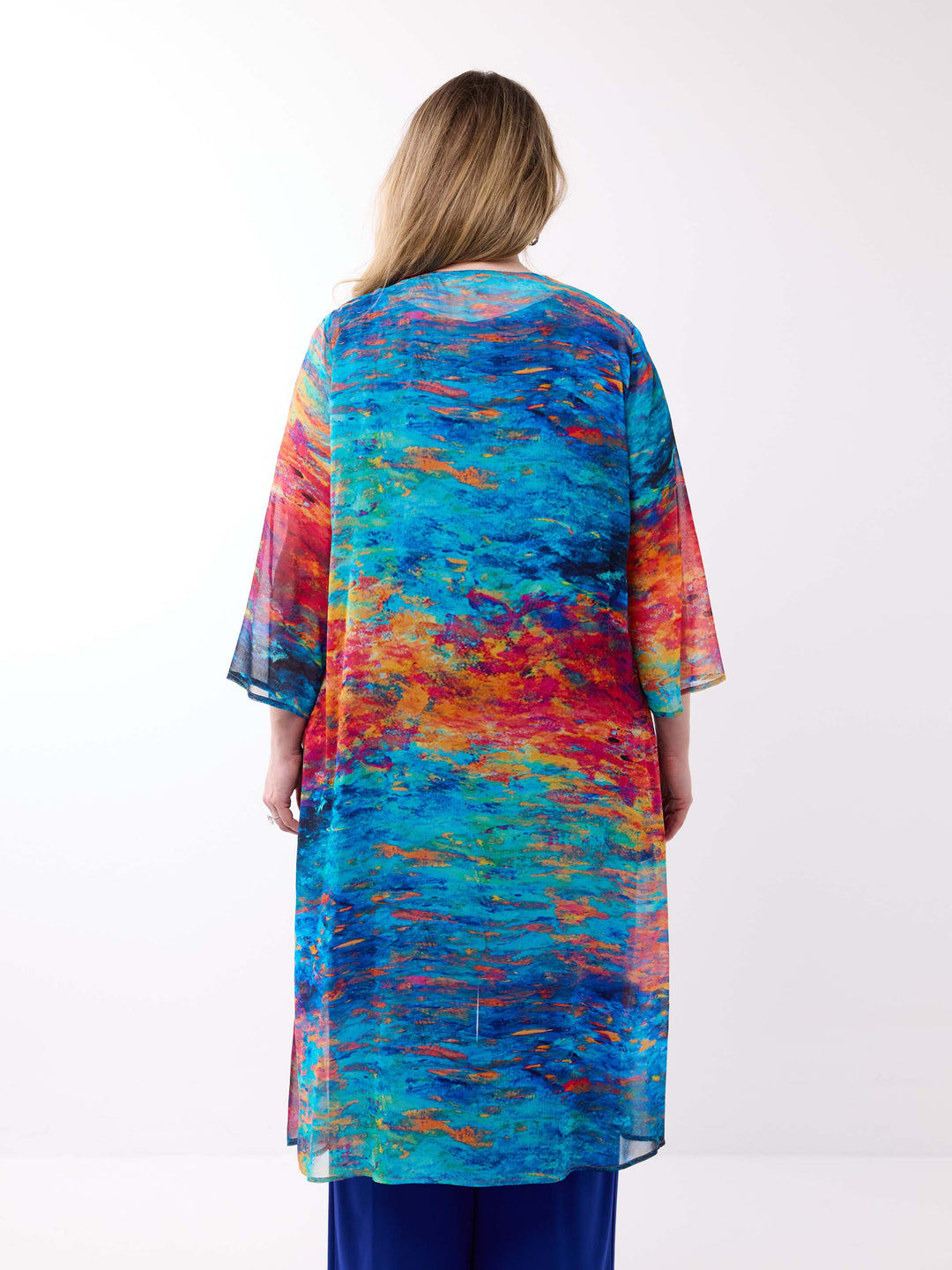 A woman viewed from behind wearing a long, flowy tunic or kaftan-style evening jacket with a vivid abstract watercolour print in blue, teal, orange, red, and yellow, paired with dark blue pants