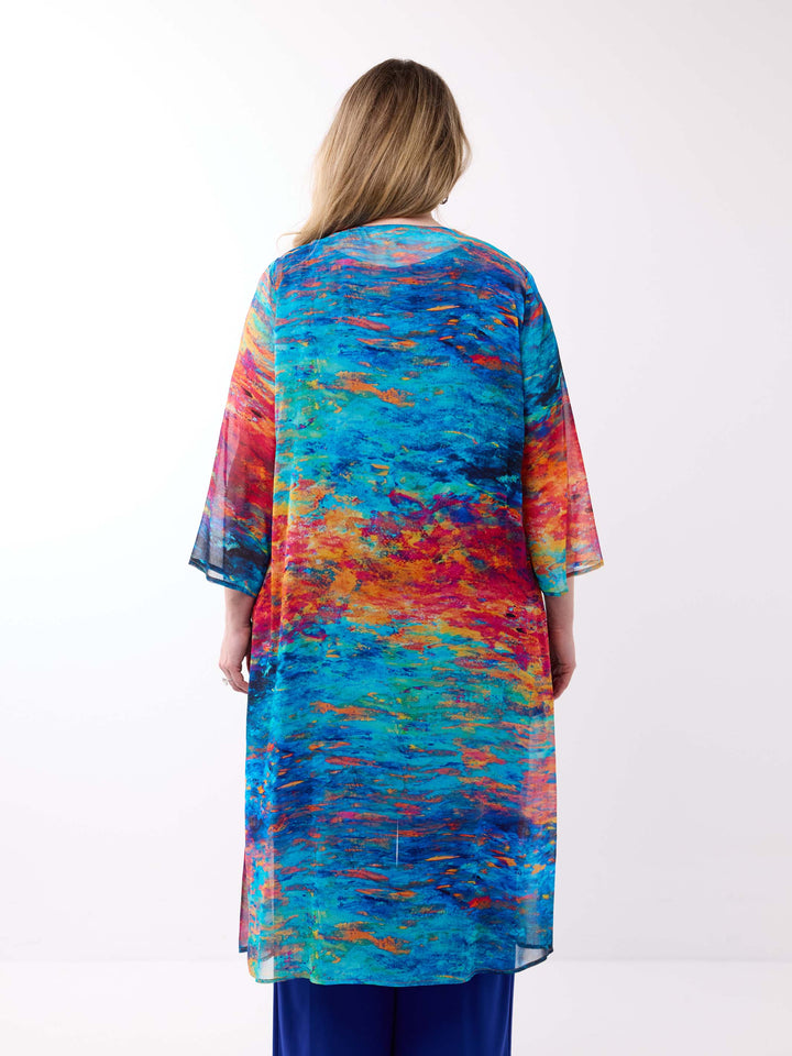 A woman viewed from behind wearing a long, flowy tunic or kaftan-style evening jacket with a vivid abstract watercolour print in blue, teal, orange, red, and yellow, paired with dark blue pants