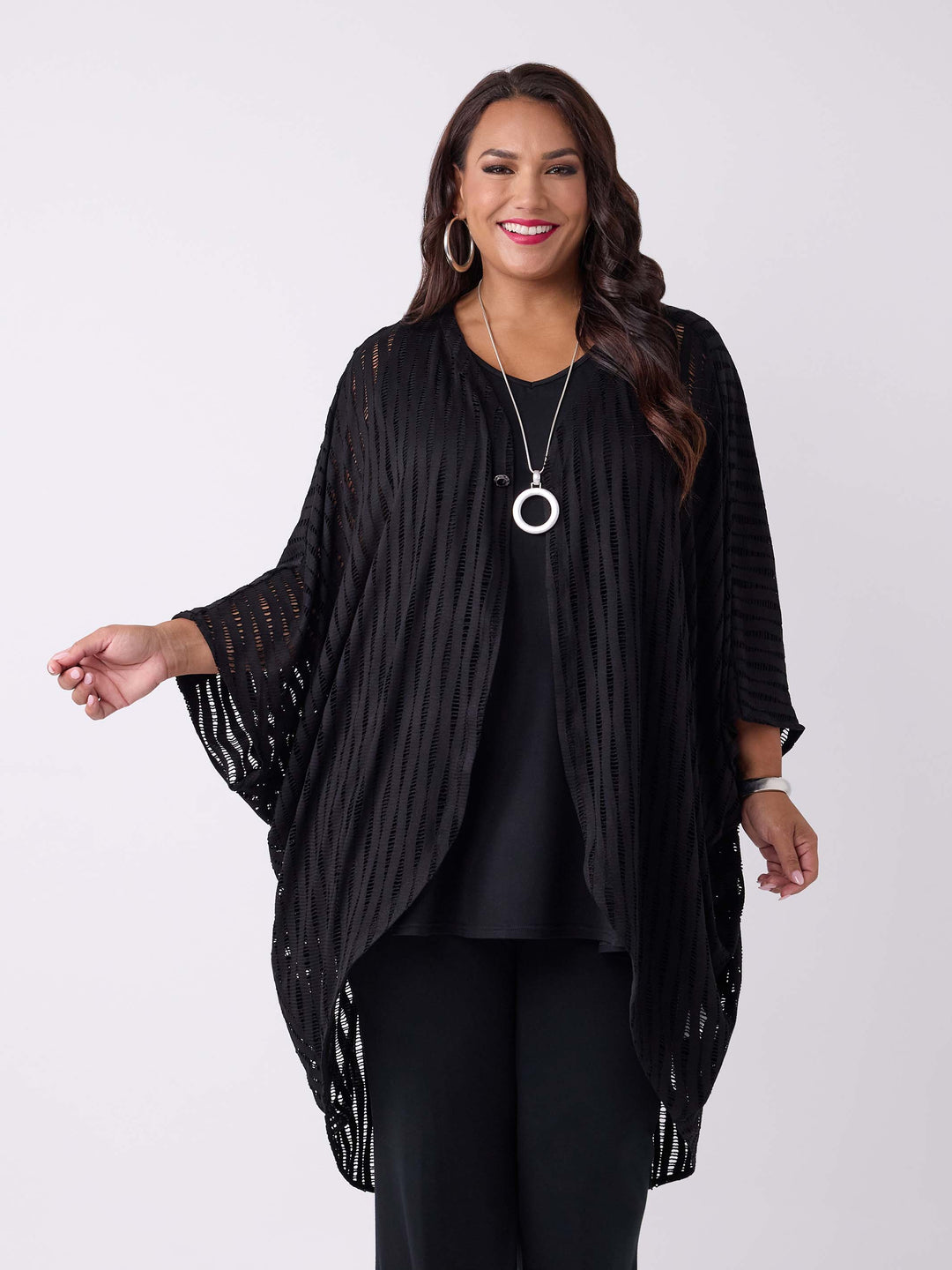 A woman wearing a black drapery longline evening jacket with ladder lace detail, styled with a black v-neck swing cami and black palazzo pant.