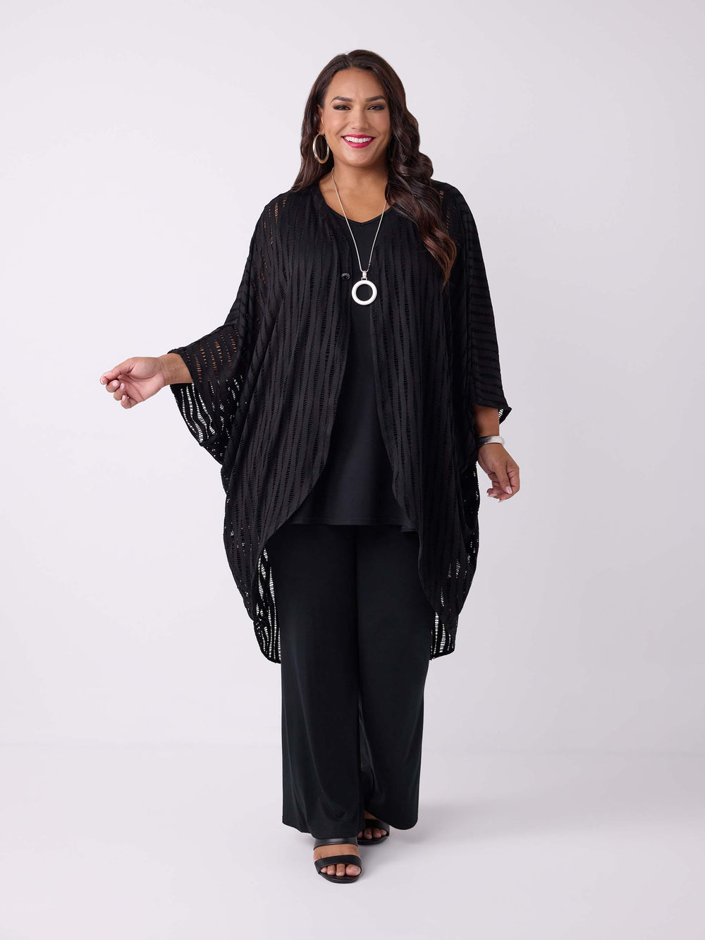 A woman wearing a black drapery longline evening jacket with ladder lace detail, styled with a black v-neck swing cami and black palazzo pant.