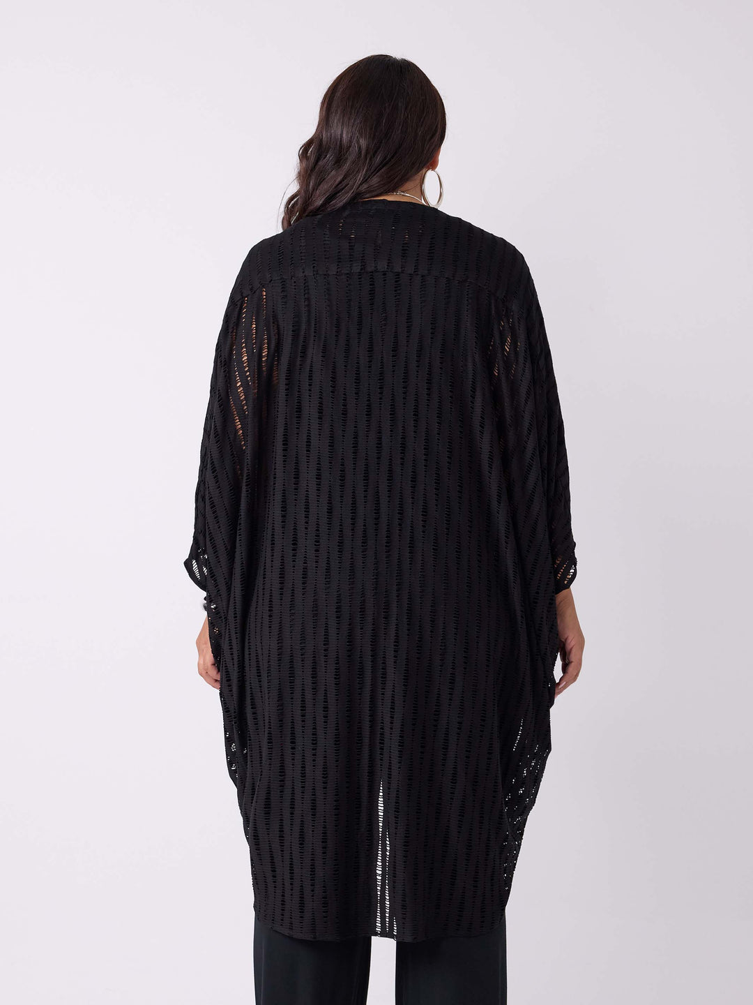 A woman viewed from the back wearing a black drapery longline evening jacket with ladder lace detail, styled with a black v-neck swing cami and black palazzo pant.