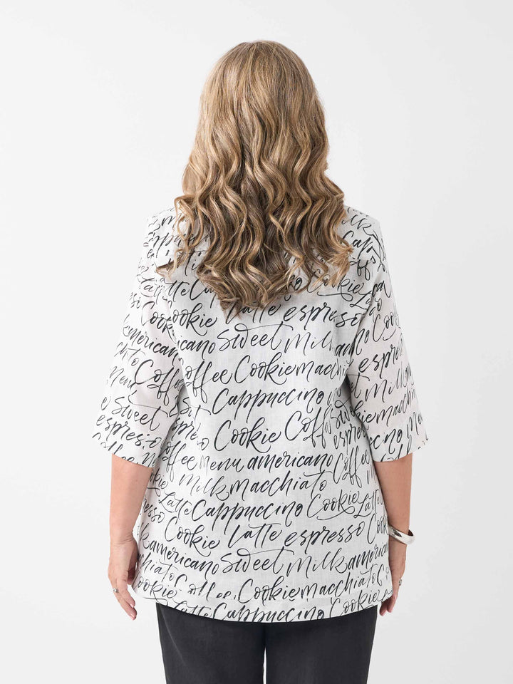 A woman viewed from the back wearing a white short-sleeve top with an all-over black script coffee-themed print, paired with black pants