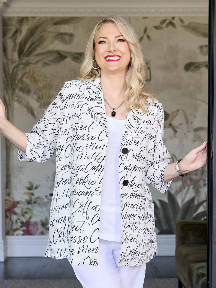 A woman stands wearing a white button-front blazer covered in black script words, over a white top and white pants.