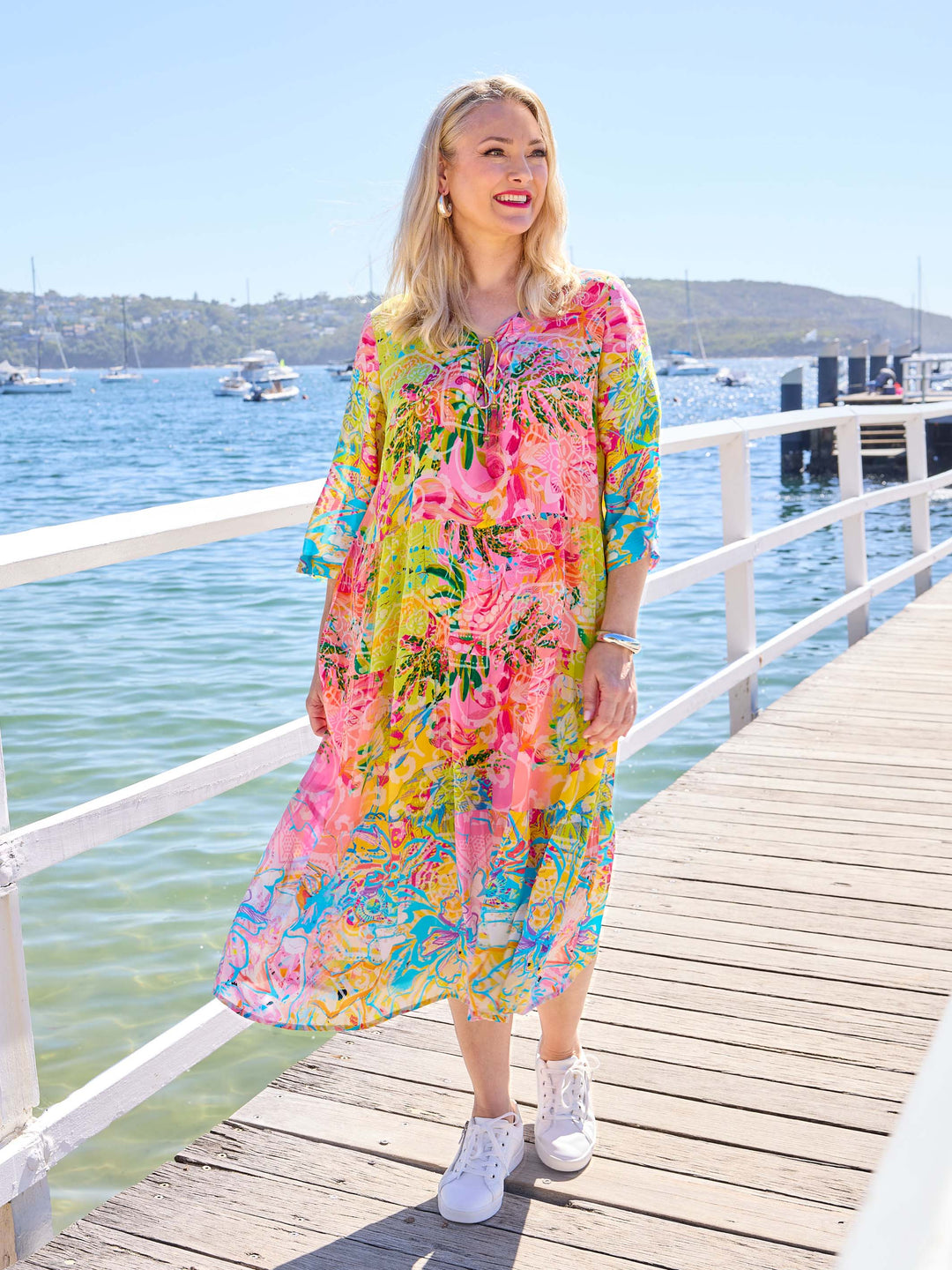 A woman wearing a bright tropical floral maxi dress with three-quarter sleeves. The dress has a vibrant multicolour print with pink, yellow, green, and blue tones.