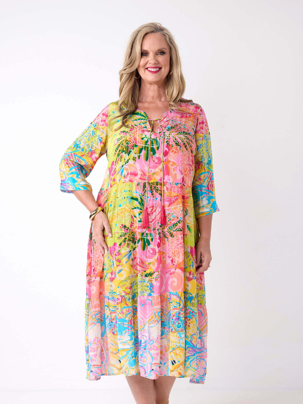 A woman wearing a bright tropical floral maxi dress with three-quarter sleeves. The dress has a vibrant multicolour print with pink, yellow, green, and blue tones.