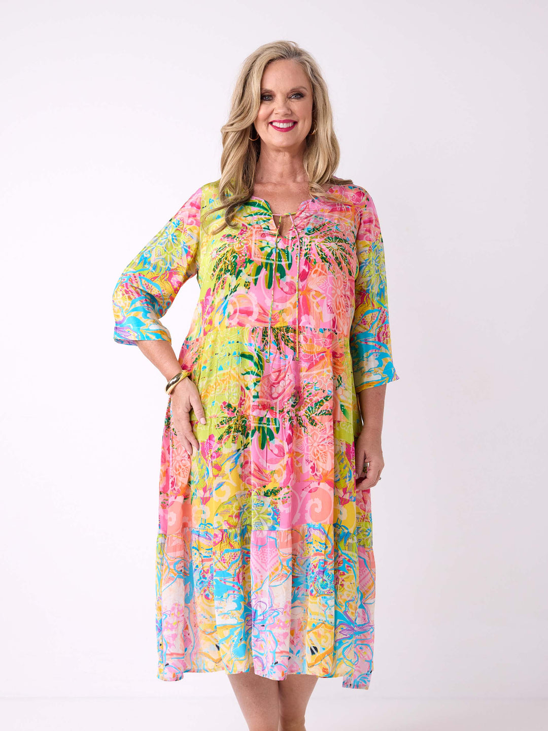 A woman wearing a bright tropical floral maxi dress with three-quarter sleeves. The dress has a vibrant multicolour print with pink, yellow, green, and blue tones.