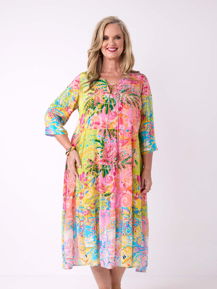 A woman wearing a bright tropical floral maxi dress with three-quarter sleeves. The dress has a vibrant multicolour print with pink, yellow, green, and blue tones.