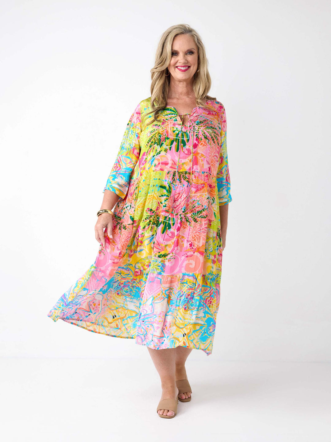 A woman wearing a bright tropical floral maxi dress with three-quarter sleeves. The dress has a vibrant multicolour print with pink, yellow, green, and blue tones.