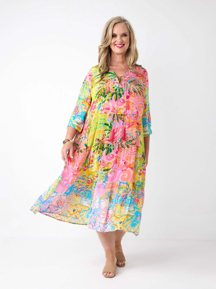 A woman wearing a bright tropical floral maxi dress with three-quarter sleeves. The dress has a vibrant multicolour print with pink, yellow, green, and blue tones.