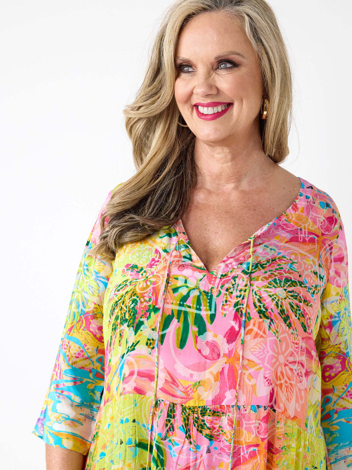 A woman wearing a bright tropical floral maxi dress with three-quarter sleeves. The dress has a vibrant multicolour print with pink, yellow, green, and blue tones.