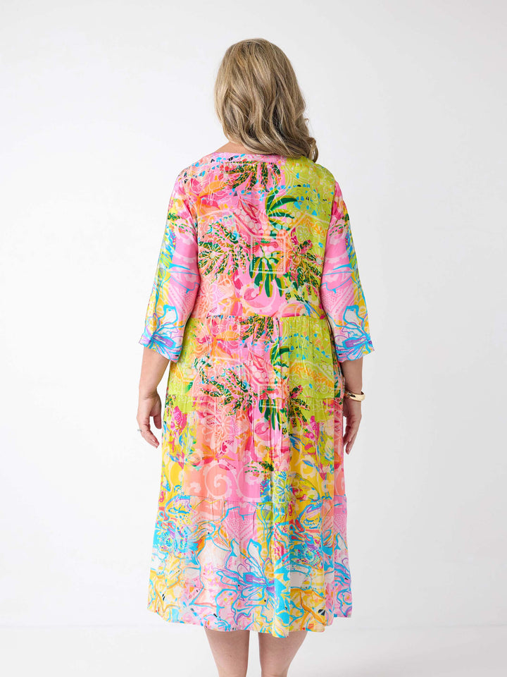 A woman wearing a bright tropical floral maxi dress with three-quarter sleeves. The dress has a vibrant multicolour print with pink, yellow, green, and blue tones.