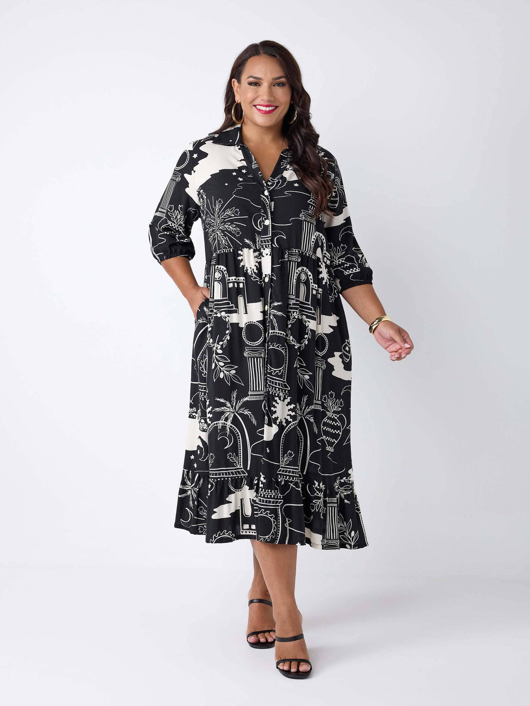 A woman wearing a black collared button through shirt midi dress with cream white roman style print, featuring ruffled hem