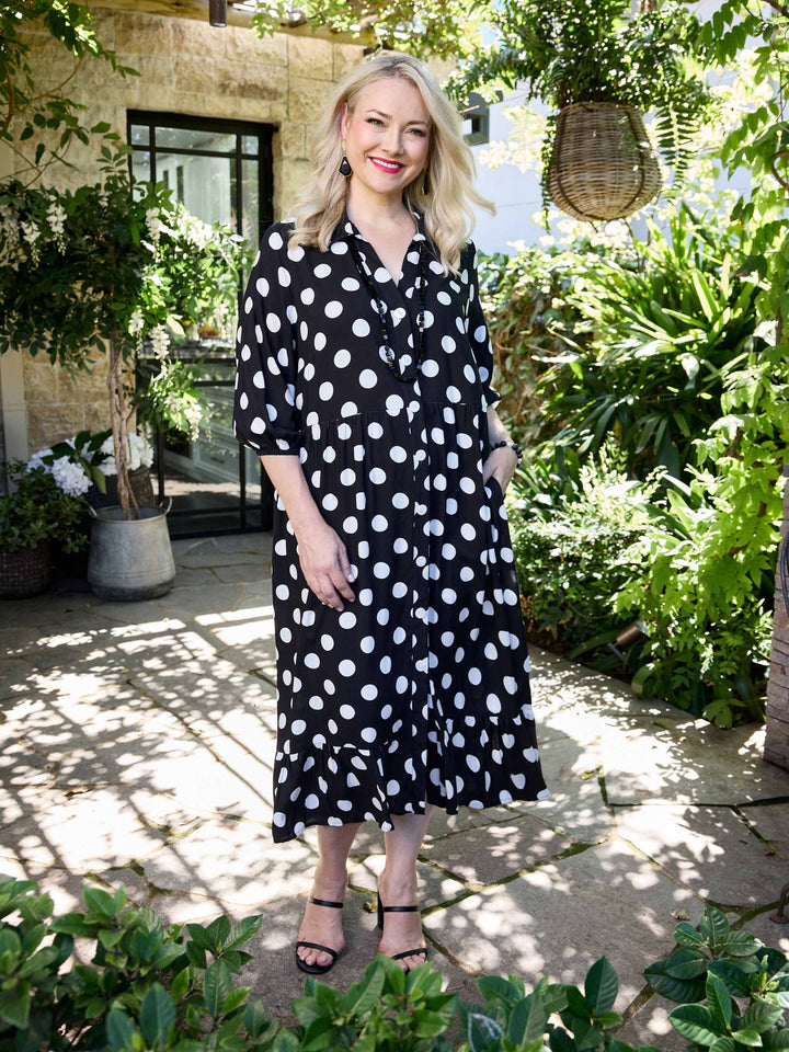 A woman wearing a black midi shirt dress with white polka dots. The dress has three-quarter sleeves, a button-up front, and a ruffled hem.