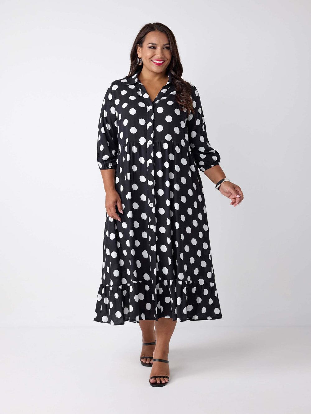 A woman wearing a black midi shirt dress with white polka dots. The dress has three-quarter sleeves, a button-up front, and a ruffled hem.
