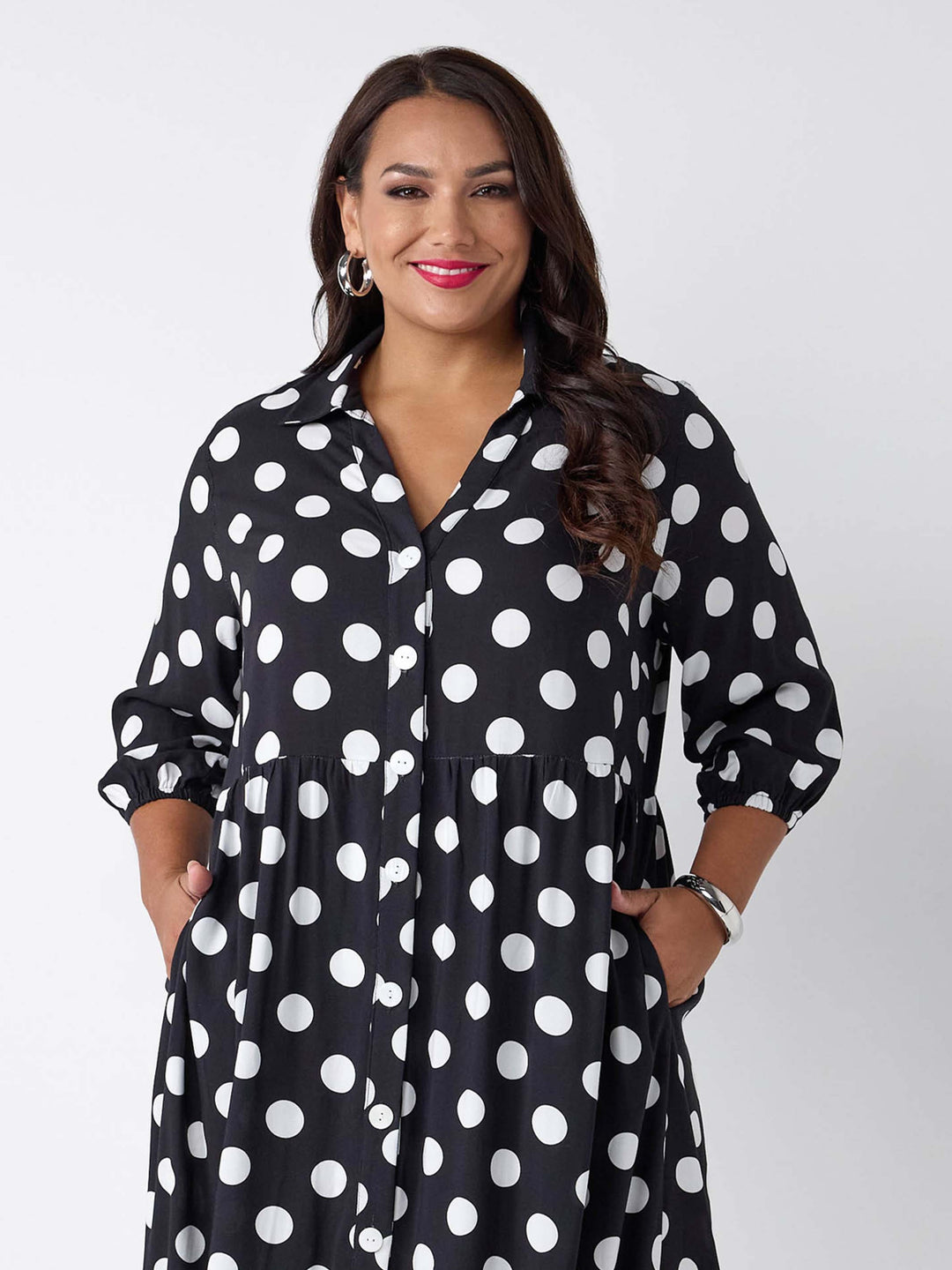A woman wearing a black button-up shirt dress with a white polka dot pattern, featuring three-quarter sleeves and side pockets
