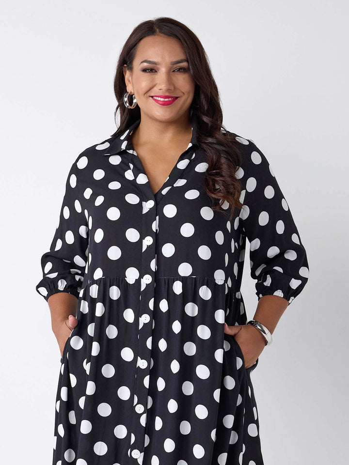 A woman wearing a black button-up shirt dress with a white polka dot pattern, featuring three-quarter sleeves and side pockets