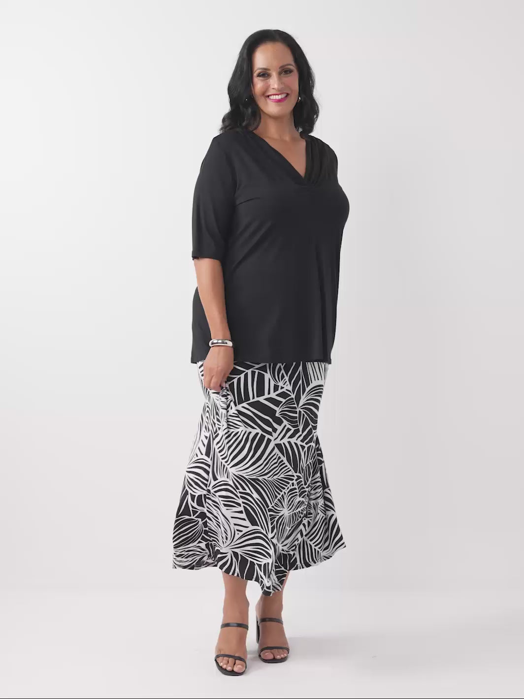 A woman wearing a black short-sleeve top with a draped cowl neckline, paired with black and white tropical palm patterned skirt