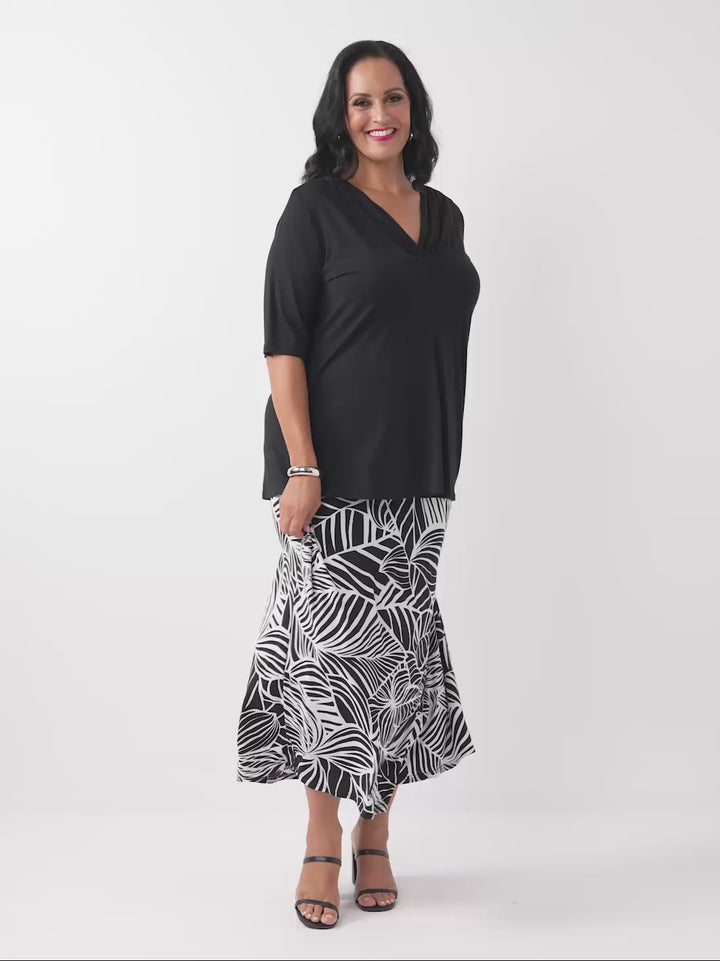 A woman wearing a black short-sleeve top with a draped cowl neckline, paired with black and white tropical palm patterned skirt