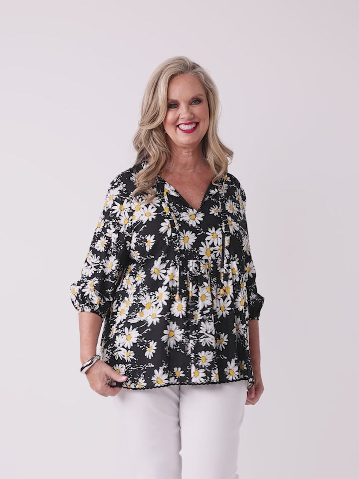 A woman wearing a black long-sleeve blouse with a white and yellow daisy floral print, paired with white pants