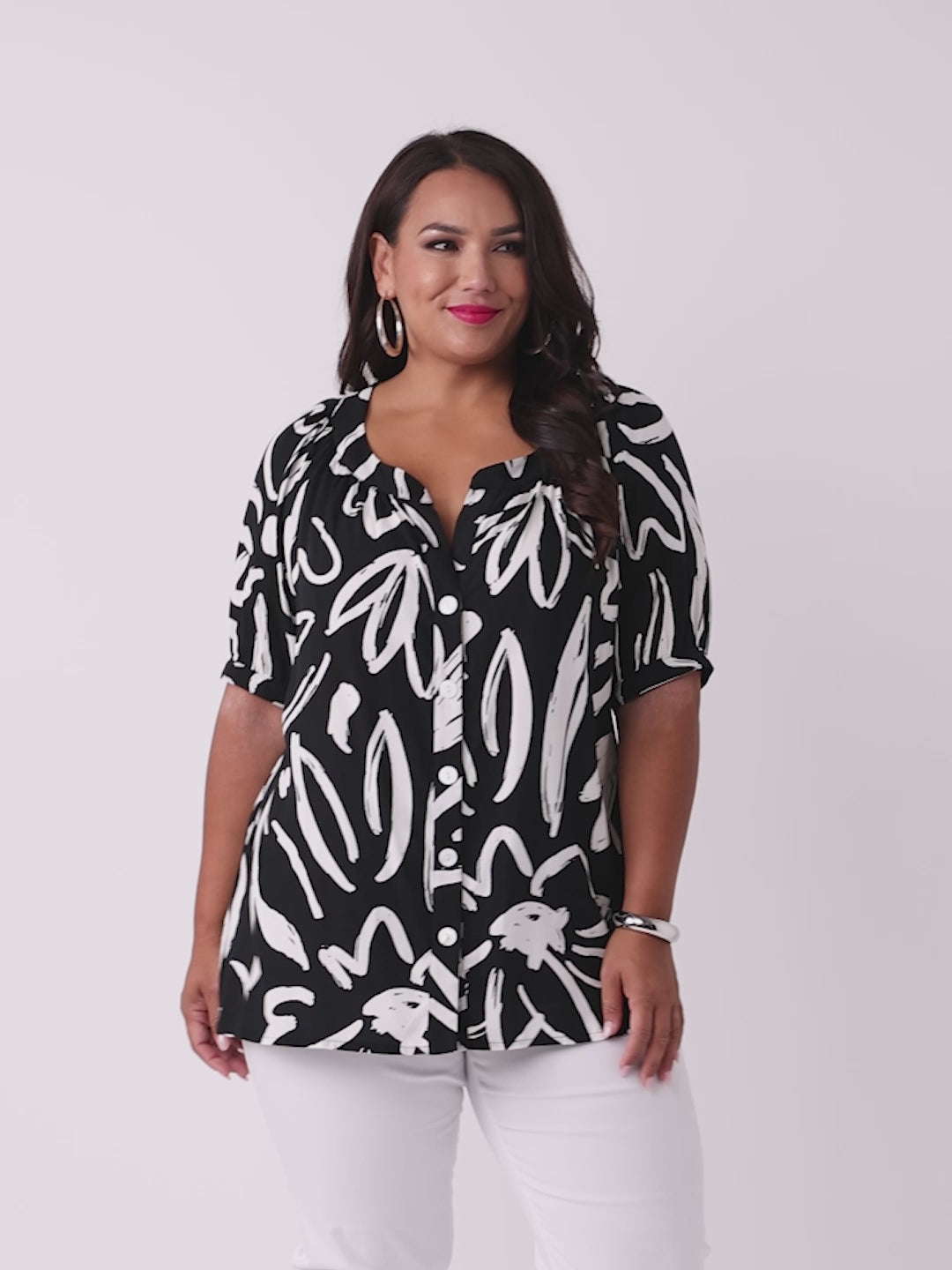 A woman wearing a black short-sleeve button-up blouse with a white abstract brushstroke print, paired with white pants