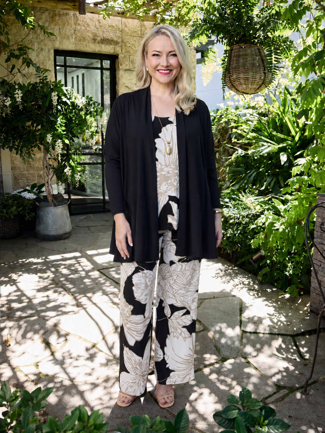 A woman stands in a garden courtyard wearing a black cardigan over a black and beige floral cami and matching palazzo pant